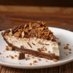 A slice of No-Bake Snickers Pie on a white plate, featuring layers of creamy peanut butter filling, chocolate, caramel, and crunchy toppings