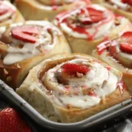 Fresh strawberry cheesecake cinnamon rolls in a baking tray with cream cheese frosting and sliced strawberries on top.