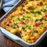 A freshly baked sausage hashbrown breakfast casserole in a white dish, topped with melted cheese, diced tomatoes, and chopped parsley.